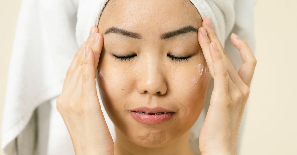 Asian woman with a towel on head applying moisturizer for skincare routine.